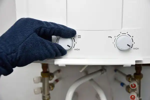 A gloved hand carefully adjusts the temperature dial on a tankless water heater panel, surrounded by visible pipes and valves, ensuring proper maintenance for efficient performance.