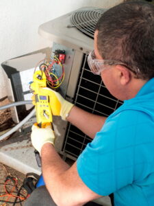 A person wearing safety glasses and gloves is using a yellow handheld device to test or repair wires connected to an HVAC unit.