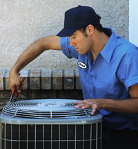 A technician in a blue uniform and cap uses a tool to service an outdoor air conditioning unit.