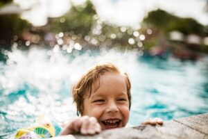 Child smiling while holding onto the edge of a swimming pool, with water splashing around them.
