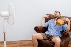 Man sitting in a brown chair, wearing a light blue shirt and holding a hat, looking hot and tired, with a white fan nearby.