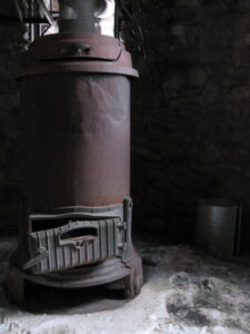 A rusty, cylindrical metal stove with a closed hatch stands on a stone floor inside a dimly lit room. A metal bucket is nearby against a stone wall.